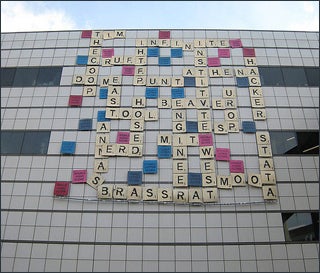 On the side of a building hang multi-colored squares, each with a letter or phrase on them, simulating a Scrabble game.