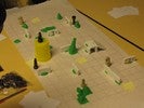 Mid-game, the board contains several structures of dominos, hills and lakes of green Play-Doh, and chess pieces with Play-Doh arms or faces. One rook is standing on its head.