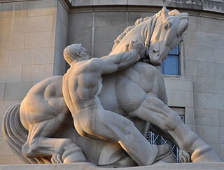 A photograph of a stone statue depicting a muscular and shirtless man pulling on the reins of an equally muscular horse.