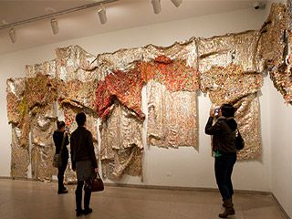 Three people stand in front of a multi-paneled drape-like sculpture made of brightly colored aluminum bottle caps and copper wire.