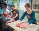 A glove-wearing student stands over a plastic water-filled containers filled with a pink, cotton-like substance. To his left, another students spreads a thin, flat, dry,pink-cotton-like substance on a table.