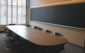 A small classroom with a large wood table surrounded by grey chairs, with chalkboards on either side.