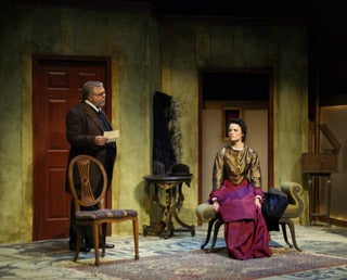 A gentleman speaks to an elegantly dressed woman seated on a chaise lounge.