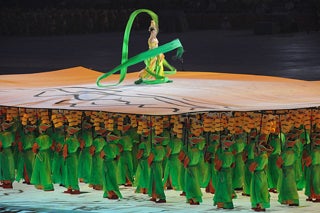 Dozens of people dressed in bright green uniforms carry poles that support a movable stage. A dancer twirling a green ribbon, stands on the stage.