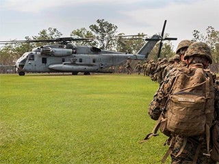 A photograph of a line of Marines wearing camouflage walking across a grass field to board a large helicopter.