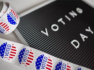 A strip of red, white, and blue stickers with the words “I Voted” lies next to an announcement board with the words “Voting Day” written upon it.