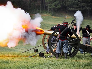 A photograph of a group of people, dressed in Civil War uniforms, standing by a cannon. The cannon has just gone off.