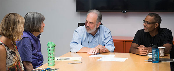 Two men sitting at the head of a table, looking toward to two women sitting on the left side of the table. One of the women is talking.