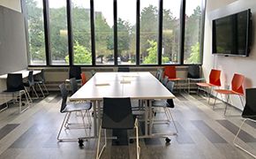 Four tables pushed together, surrounded by black and orange chairs. Wall of windows. Chalkboard on the left. Large flat screen monitor on the right.